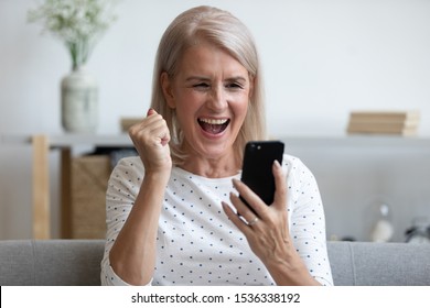 Overjoyed mature old woman hold phone looking at screen feel excited about mobile online app bid win, happy middle aged lady winner celebrate victory success read message good news in cell at home