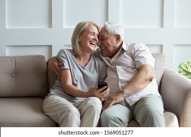 Overjoyed Mature Retired Family Spouse Watching Funny Photos On Mobile Phone, Sitting On Sofa. Happy Bonding Older Married Couple Relaxing On Couch, Laughing, Enjoying Leisure Time Together At Home.