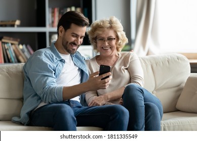 Overjoyed Mature Woman Wearing Glasses And Adult Son Using Smartphone Together, Looking At Screen, Elderly Mother And Young Man Hugging, Having Fun With Phone, Watching Funny Video, Shopping Online