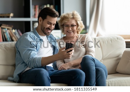 Overjoyed mature woman wearing glasses and adult son using smartphone together, looking at screen, elderly mother and young man hugging, having fun with phone, watching funny video, shopping online