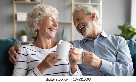 Overjoyed Middle Aged Retired Married Couple Relaxing On Comfortable Sofa, Enjoying Communicating While Drinking Hot Morning Coffee Together At Home, Laughing At Funny Joke, Family Relations Concept.