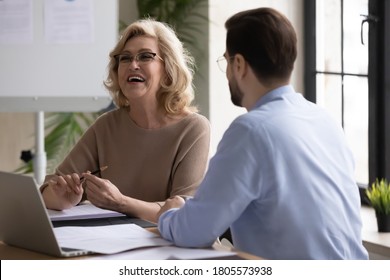 Overjoyed middle aged senior female leader having fun, enjoying informal conversation with young male colleague at office meeting. Happy mature mentor laughing at funny joke with partner indoors.
