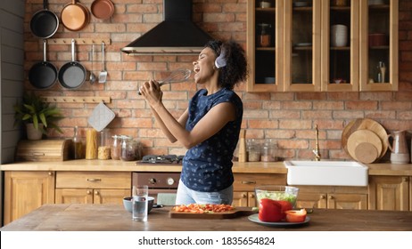 Overjoyed millennial African American woman have fun cooking healthy tasty breakfast in home kitchen. Happy playful young biracial girl sing entertain enjoy good morning prepare food at countertop.