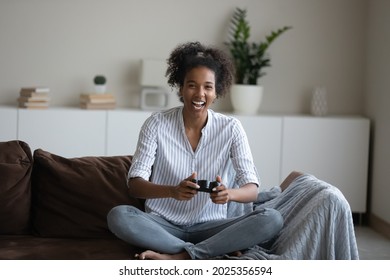 Overjoyed Millennial African American Woman Sit Relax On Sofa In Living Room Have Fun Play Video Computer Game. Happy Smiling Young Ethnic Female Gamer Enjoy Weekend With Virtual Reality Videogame.