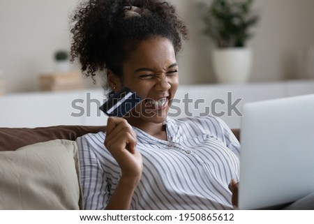 Overjoyed millennial African American woman have fun shopping online with credit card on laptop. Excited young biracial female buy pay on internet on computer at home use good banking secure system.