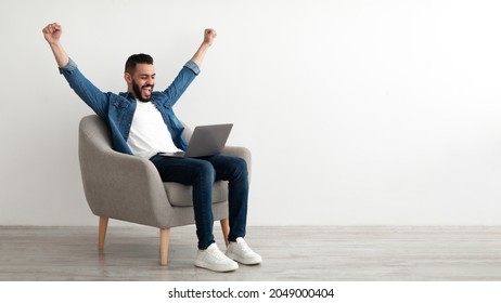 Overjoyed millennial Arab man sitting in armchair with laptop, celebrating online win or work success against white studio wall, copy space. Young Eastern guy enjoying big sale in web store, banner - Shutterstock ID 2049000404