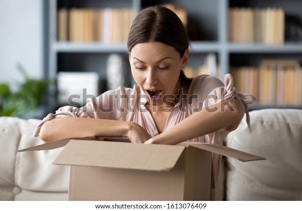 Overjoyed millennial girl sit on sofa in living room unpack cardboard