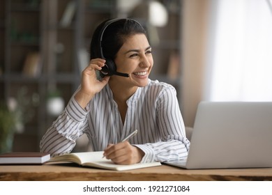 Overjoyed millennial Indian woman in earphones study online on laptop take notes. Happy young mixed race female in headphones work distant on computer. Education, virtual event concept.