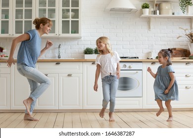 Overjoyed Millennial Mom Or Nanny Dancing With Little Girls Children In White Modern Kitchen, Happy Young Caucasian Mother Have Fun Involved In Activity Together With Small Daughters At Home