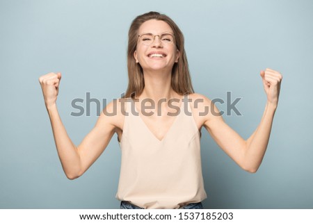 Overjoyed millennial woman in glasses isolated on blue studio background feel euphoric with good news, excited young Caucasian female winner raise hands celebrate victory or success, reward concept
