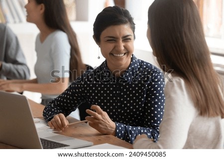 Overjoyed multiracial millennial female students sit at desk in classroom laugh working on educational project together, smiling multiethnic girls have fun joke brainstorming cooperating at lesson