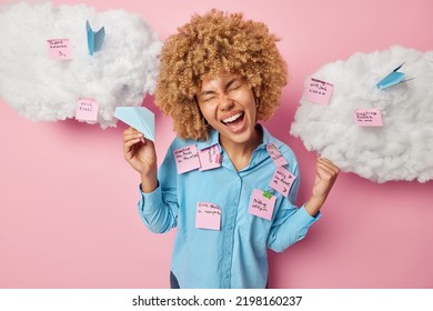 Overjoyed Multitasking Female Model Makes Fist Bump Celebrates Victory Holds Paper Aeroplane Glad To Finish One Task From Variety Writes Down Notes On Stickers Dressed Casually Isolated On Pink Wall