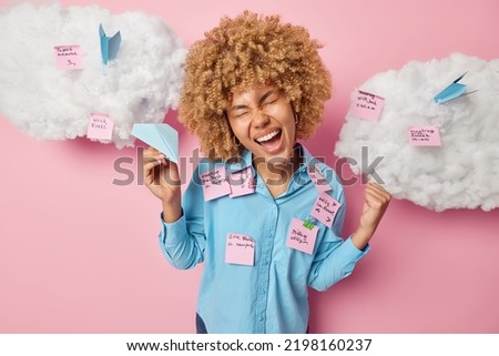 Overjoyed multitasking female model makes fist bump celebrates victory holds paper aeroplane glad to finish one task from variety writes down notes on stickers dressed casually isolated on pink wall
