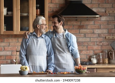 Overjoyed Older 60s Caucasian Father Have Fun Cooking Together With Adult Son In Kitchen. Smiling Young Man Child Enjoy Preparing Healthy Salad For Dinner With Mature Dad At Home. Hobby Concept.