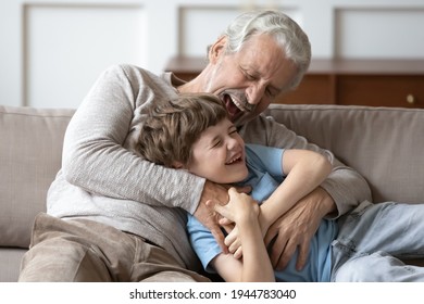 Overjoyed older Caucasian grandfather relax in living room with smiling little grandson tickle and giggle. Smiling mature granddad feel playful have fun play with small 8s boy child at home together.