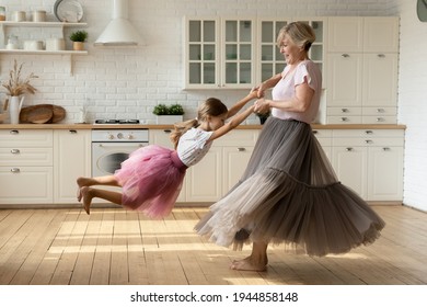 Overjoyed Optimistic Mature 60s Grandmother And Little 8s Granddaughter Wear Princess Skirts Dresses Dance Together In Kitchen. Happy Elderly Granny Have Fun Play Enjoy Weekend With Small Grandchild.