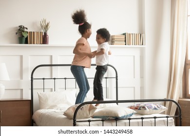 Overjoyed Playful African American Small Brother And Sister Laugh Playing Jumping On Bed At Home, Excited Happy Black Boy And Girl Kids Feel Energetic Have Fun Engaged In Funny Activity In Bedroom