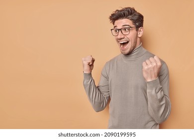 Overjoyed positive man clenches fists with triumph rejoices excellent news exclaims from happiness cannot believe in success wears casual jumper and spectacles gesture actively poses indoor.
