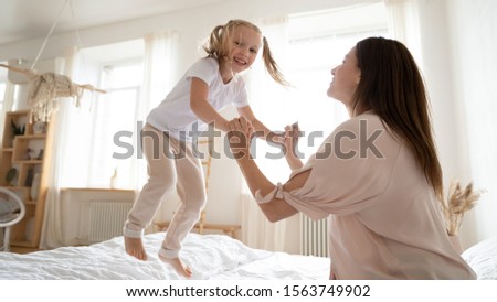 Overjoyed preschool adorable daughter jumping on bed, holding happy mommys hands, looking at camera. Joyful family of two having fun in bedroom, enjoying active weekend morning together at home.