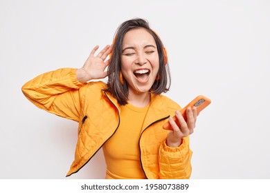 Overjoyed pretty Asian teenage girl got carried away with music holds smartphone enjoys favorite playlist via stereo headphones has relaxed carefree expression sings along wears orange jacket