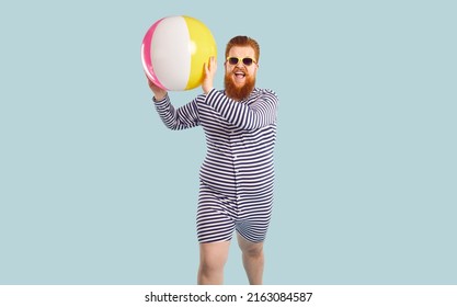 Overjoyed redhead fat man in stripy swimsuit isolated on grey blue studio background play with ball. Funny red-haired guy enjoy summer vacation or holidays. Summertime activity and fun.