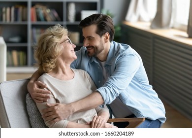 Overjoyed senior 70s mother sit in chair talk laugh enjoy family weekend with grown-up son at home, happy mature mom and adult man child embrace hug having tender close moment together