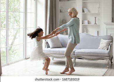 Overjoyed Senior Grandmother Dancing Entertaining With Little Granddaughter In Living Room, Happy Grandparent Have Fun Engaged In Funny Family Activity With Small Grandchild At Home Together