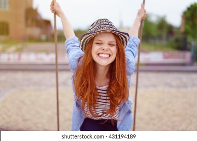 Overjoyed single red haired young woman in hat and blue denim shirt with rolled up sleeves sitting on swing at urban playground