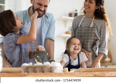 Overjoyed Small Children Siblings Spreading Flour On Laughing Parents, Enjoying Cooking Homemade Pastry Together In Kitchen. Happy Married Couple Involved In Funny Family Activity With Little Kids.