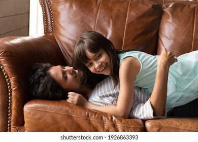 Overjoyed small Hispanic girl lying relaxing on sofa playing with young mother at home on family weekend. Excited little Latino child and mom have fun together engaged in funny game rest on couch.