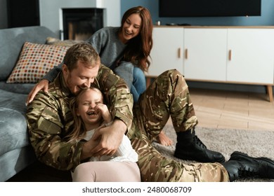 Overjoyed soldier tickling small daughter while sitting at home at the floor. Happy woman bonding to her family