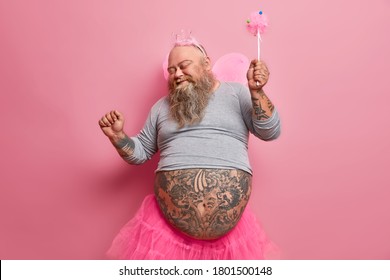 Overjoyed stout man in fairy costume dances carefree, has fun and foolishes around, organises holiday for children, plays princess, moves against pink background. Happiness, entertainment concept