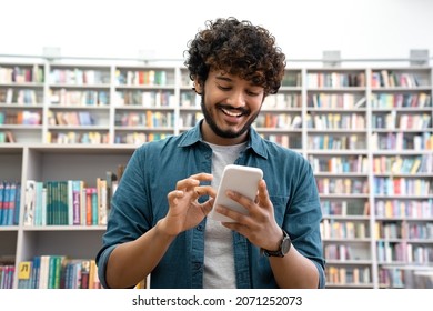 Overjoyed Successful Young Indian Student Chatting Social Media On Smartphone In Library. Happy Successful Teen In Casual Denim Shirt Shared Success In Network After Passing The Exam In Reading Room