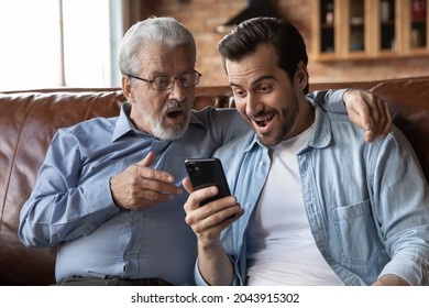 Overjoyed Surprised Two Generations Of Men Looking At Phone Screen, Excited Senior Father And Grown-up Son With Open Mouths Reading Unbelievable Good News In Message, Sitting On Couch At Home