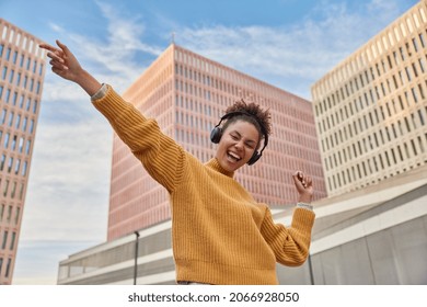 Overjoyed teenage girl feels cool dances with rhythm of music wears stereo headphones wears casual yellow jumper poses against urban bckground among skyscrapers. Hobby and lifestyle concept. - Powered by Shutterstock