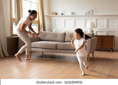 Overjoyed Vietnamese Young Mom Have Fun Run Play With Cute Smiling Biracial Daughter In Living Room, Happy Asian Mother Or Nanny Involved In Family Activity With Small Ethnic Girl Child At Home