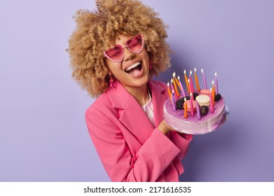 Overjoyed woman with curly hair laughs out happily wears trendy heart shaped sunglasses and formal jacket holds big tasty cake celebrates anniversary enjoys party time isolated over purple background