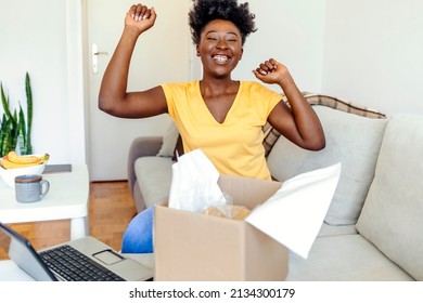 Overjoyed woman received unpacking cardboard box, sitting on the sofa in living room. Female buyer excited about her online shopping purchase. Excited black lady unboxing parcel after online shopping.