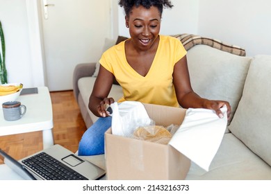 Overjoyed woman received unpacking cardboard box, sitting on the sofa in living room. Female buyer excited about her online shopping purchase. Excited black lady unboxing parcel after online shopping.