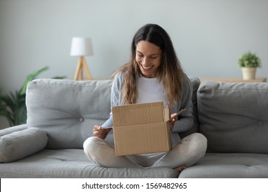 Overjoyed woman seated on sofa hold on lap small cardboard box open parcel client feels satisfied bought goods in internet, happy addressee received package from friend, quick delivery service concept - Shutterstock ID 1569482965