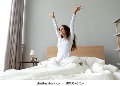 Overjoyed woman wearing pajama stretching hands after awakening, sitting in cozy bad, happy smiling attractive young female starting new day, enjoying morning after healthy sleep, doing exercises - Shutterstock ID 1890676714
