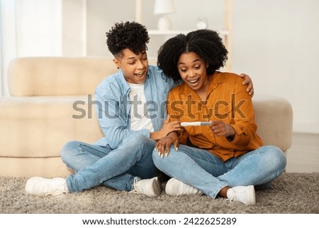 An overjoyed young african american couple sits on the floor with a positive pregnancy test, sharing an exhilarating moment of joy as they react to the good news in a cozy living room