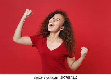 Overjoyed young african american woman girl in casual t-shirt posing isolated on red background studio portrait. People emotions lifestyle concept. Mock up copy space. Clenching fists like winner - Shutterstock ID 2077894210