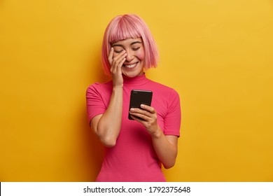 Overjoyed young Asian lady has bob hairstyle, laughs sincerely at message in social network, enjoys mobile communication, reads funny news, wears casual pink t shirt, has rosy hair with fringe