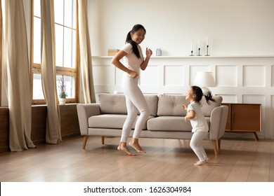 Overjoyed Young Asian Mom And Cute Little Biracial Daughter Have Fun Dancing In Living Room Together, Happy Millennial Mother Play Involved In Funny Activity With Small Vietnamese Girl Child At Home