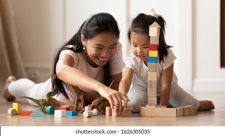 Overjoyed young Asian mom and little biracial daughter lie on warm home floor construct with building bricks together, happy millennial Vietnamese mother have fun play with small girl child