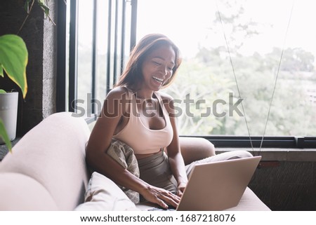 Overjoyed young attractive ethnic female in casual clothes laughing and using laptop while sitting on comfortable sofa in modern flat