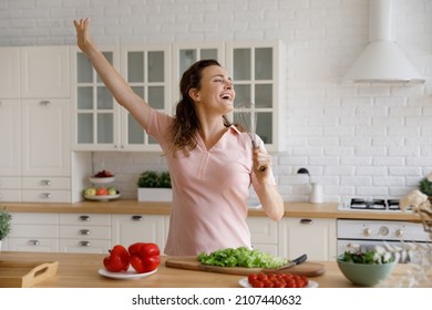 Overjoyed young beautiful woman singing favorite song in whisk as microphone, having fun cooking healthy vegetarian food alone in modern kitchen, enjoying domestic culinary hobby activity on weekend.