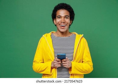 Overjoyed Young Black Curly Man 20s Years Old Wears Yellow Waterproof Raincoat Outerwear Hold In Hand Use Mobile Cell Phone Typing Browsing Chatting Isolated On Plain Green Background Studio Portrait