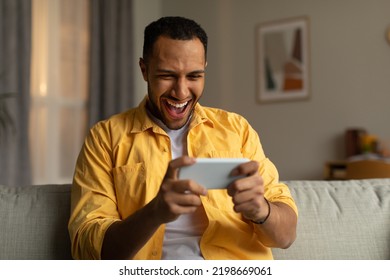 Overjoyed Young Black Man With Smartphone Feeling Excited Over Winning Lottery Or Casino Bet Online, Sitting On Couch At Home. Emotional African American Guy Playing Video Game, Gambling Indoors